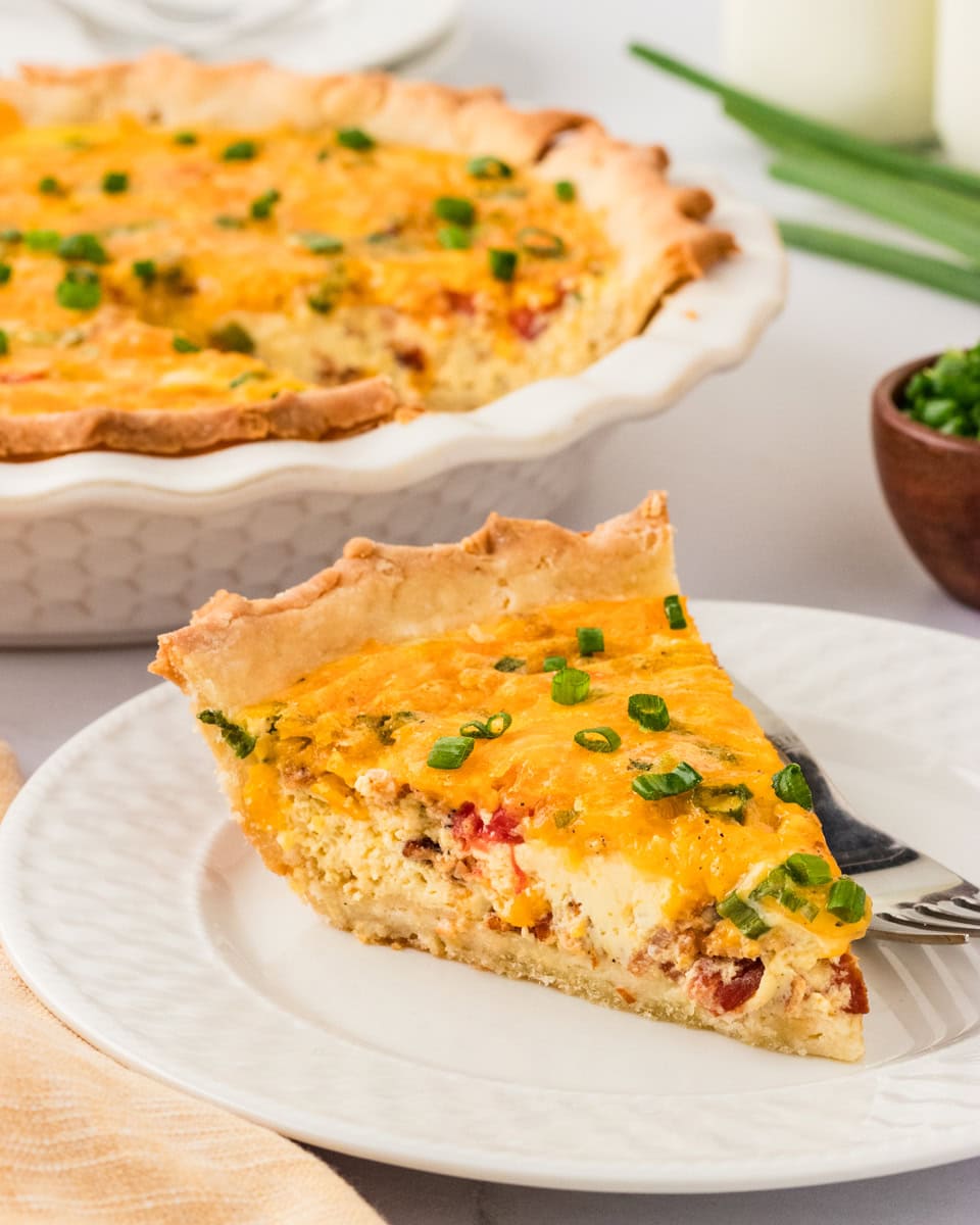 A slice of golden-brown gluten-free quiche on a white plate, garnished with green onions, with the remaining quiche in the background.