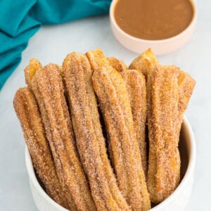 A bowl of gluten-free churros coated in cinnamon sugar, served with a caramel dipping sauce in the background.