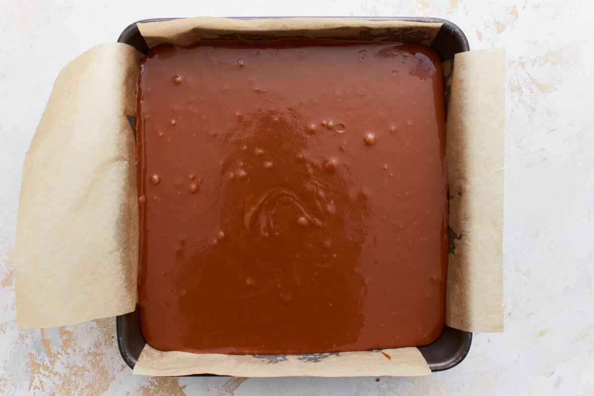 A parchment-lined baking pan filled with a smooth layer of caramel, setting atop the baked shortbread base.
