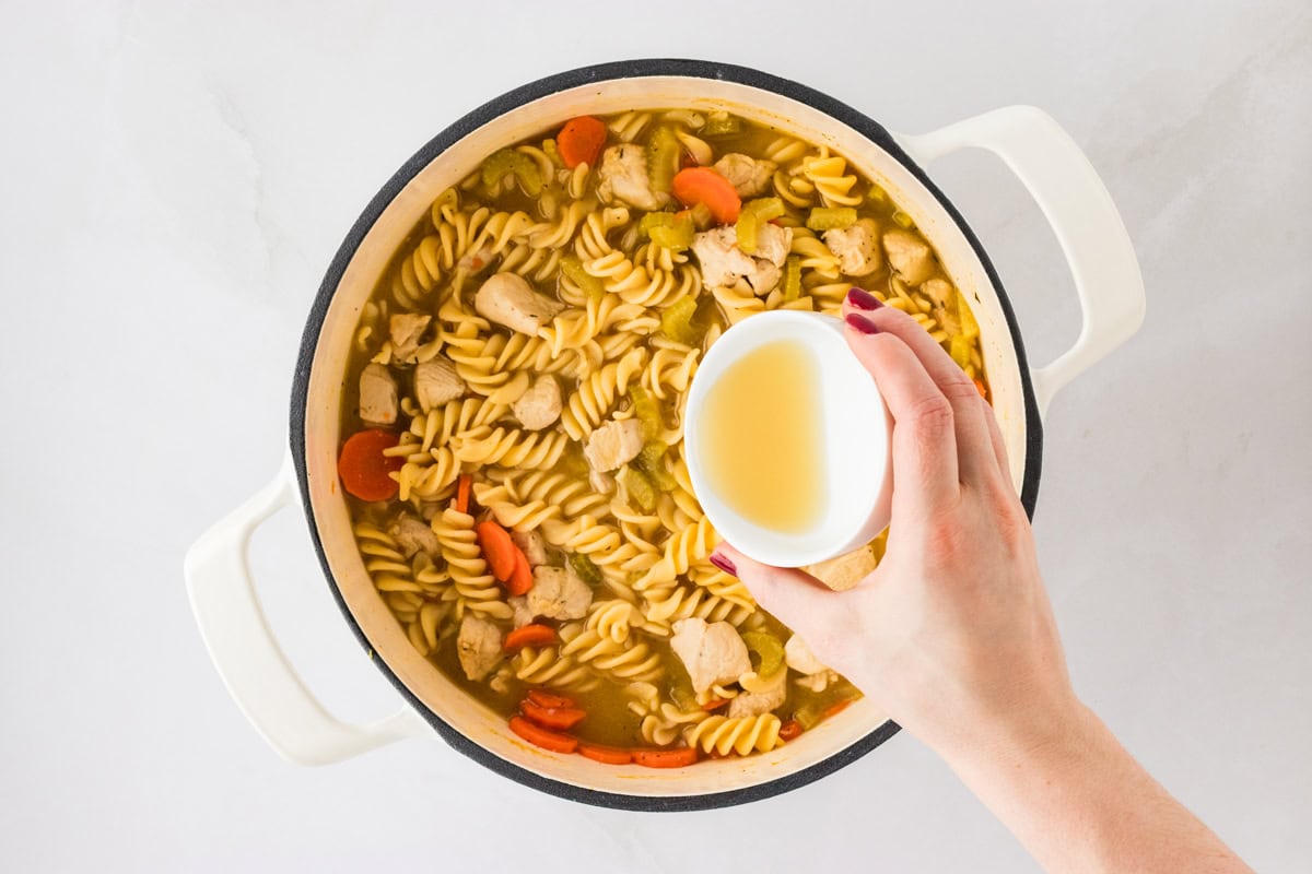 A pot of fully cooked gluten-free chicken noodle soup with vibrant vegetables, spiral noodles, and chicken chunks, while someone adds apple cider vinegar to enhance the flavor.