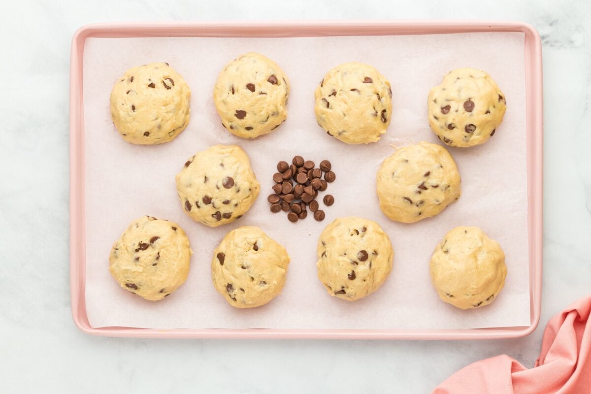 Unbaked gluten-free cookie dough balls with chocolate chips, arranged on a parchment-lined pink baking sheet.