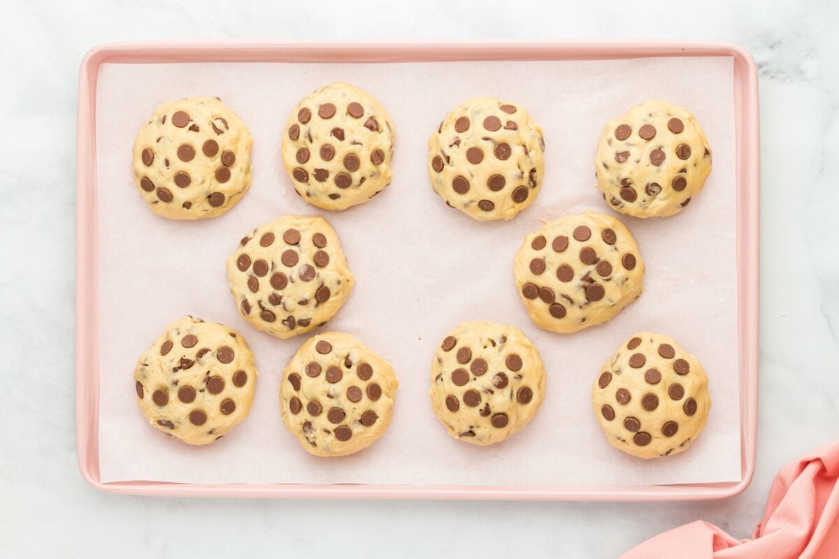 Pre-baked gluten-free Crumbl cookies with extra chocolate chips pressed into the tops, arranged neatly on a parchment-lined baking sheet.