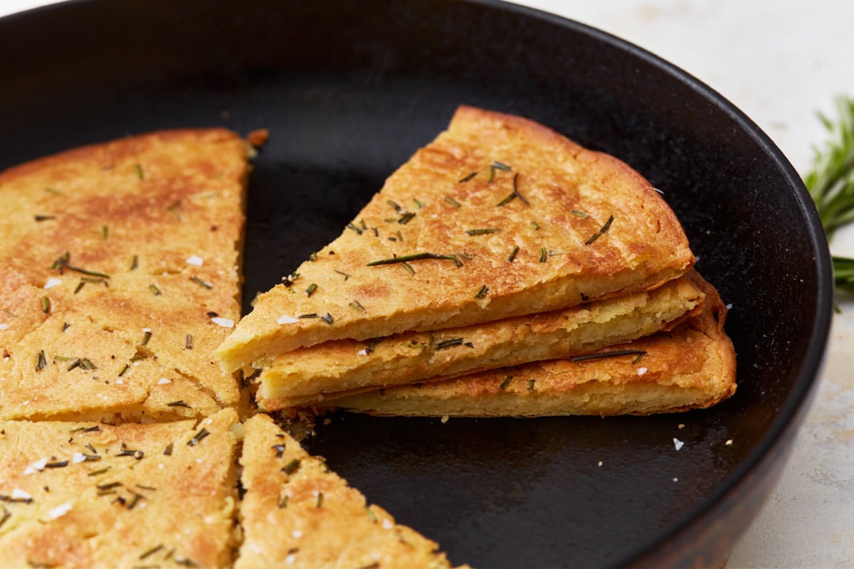 A side view of sliced farinata stacked in a cast iron skillet, showcasing its texture and golden crust. Topped with fresh rosemary and sea salt.