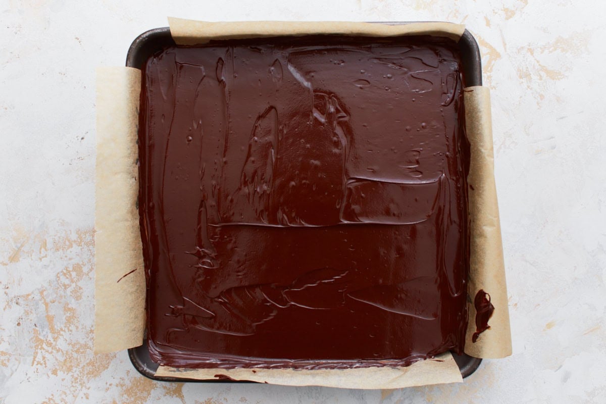 A parchment-lined baking pan with a layer of smooth melted chocolate spread over the caramel, completing the top layer.