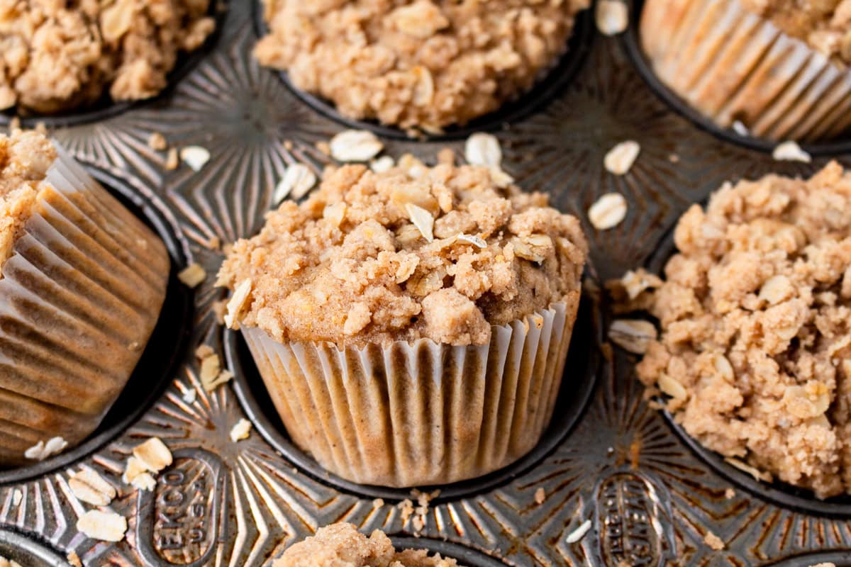 Close-up of a single banana oatmeal muffin in a muffin tin, showcasing its golden-brown crumbly oat streusel topping with scattered oats around.