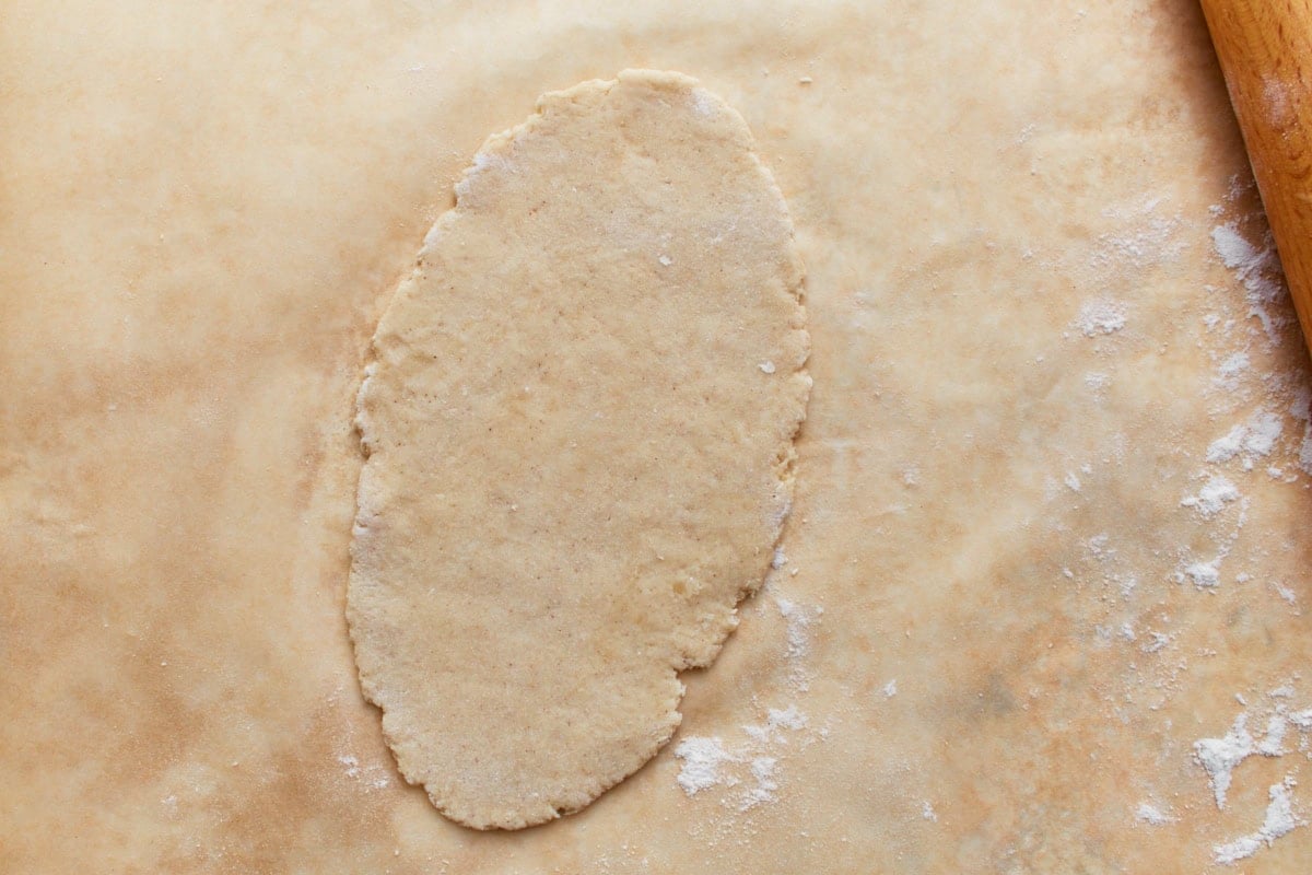 A single gluten-free naan dough ball rolled out into an oval shape on a floured surface with a rolling pin nearby.