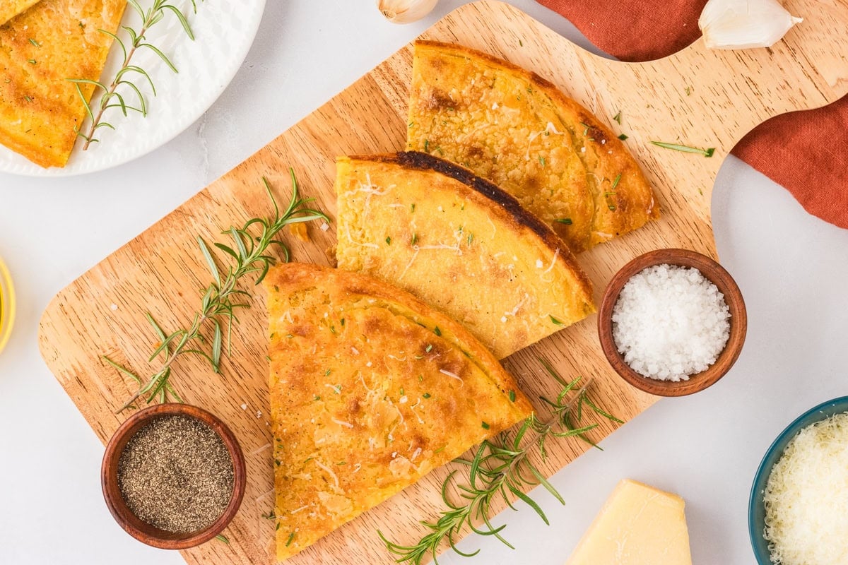 Triangular slices of socca flatbread arranged on a wooden cutting board, garnished with rosemary, salt, and pepper, with Parmesan and garlic nearby for serving.