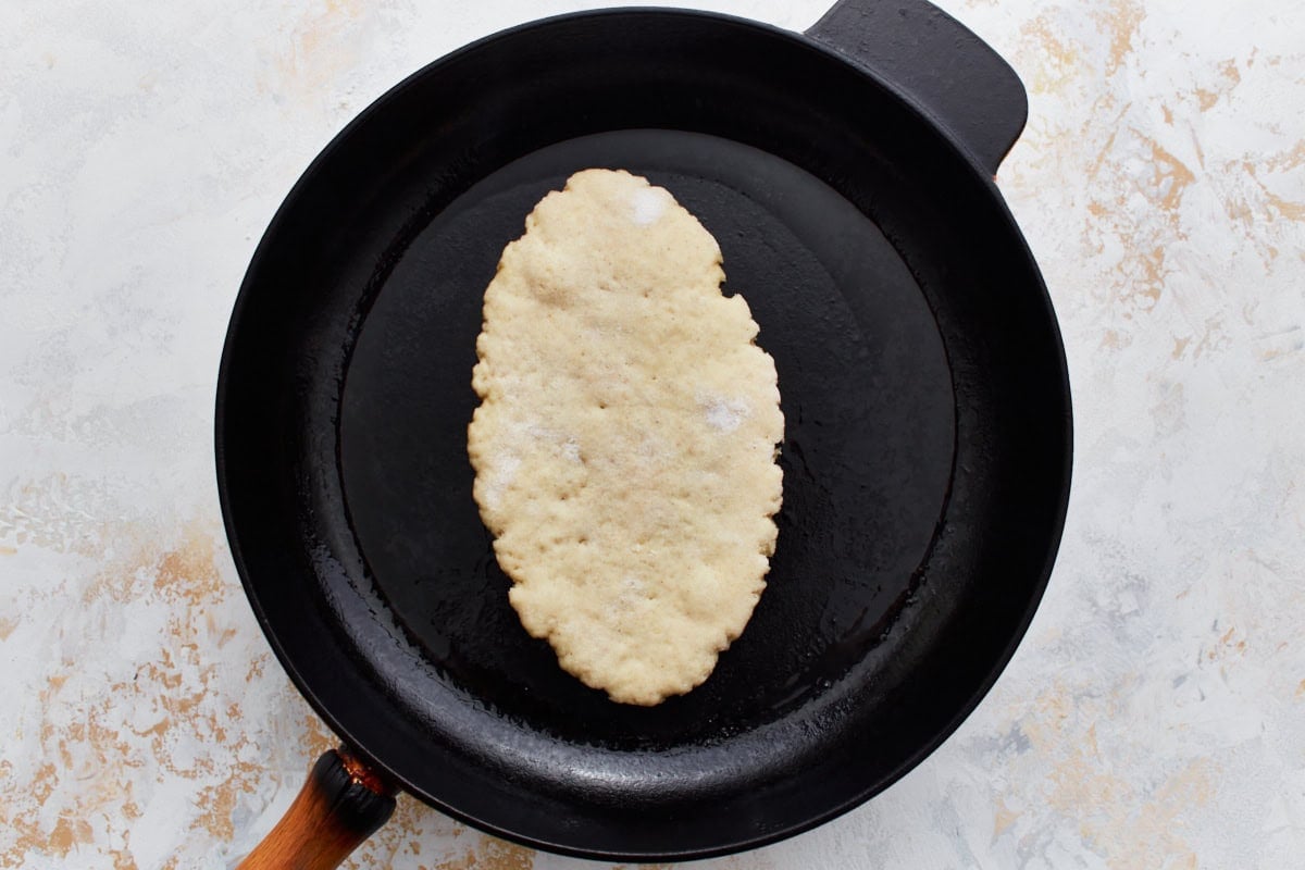 A piece of raw gluten-free naan dough cooking in a hot cast-iron skillet.