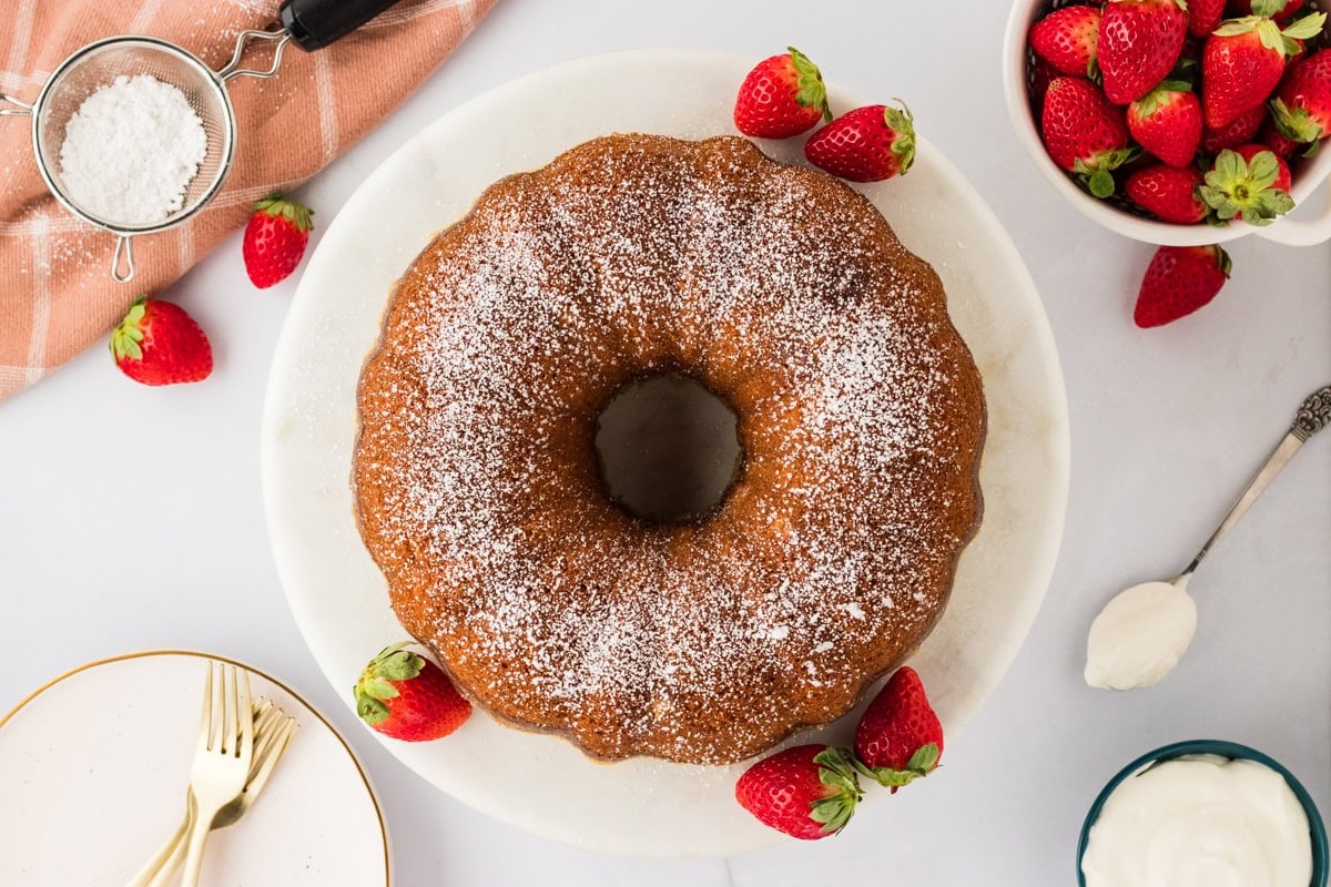 A fully prepared Gluten-Free Kentucky Butter Pound Cake dusted with powdered sugar on a white marble platter, surrounded by fresh strawberries and serving utensils.