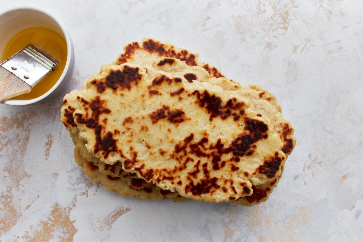 A stack of freshly cooked gluten-free naan bread brushed with melted butter next to a small bowl of melted butter with a brush.