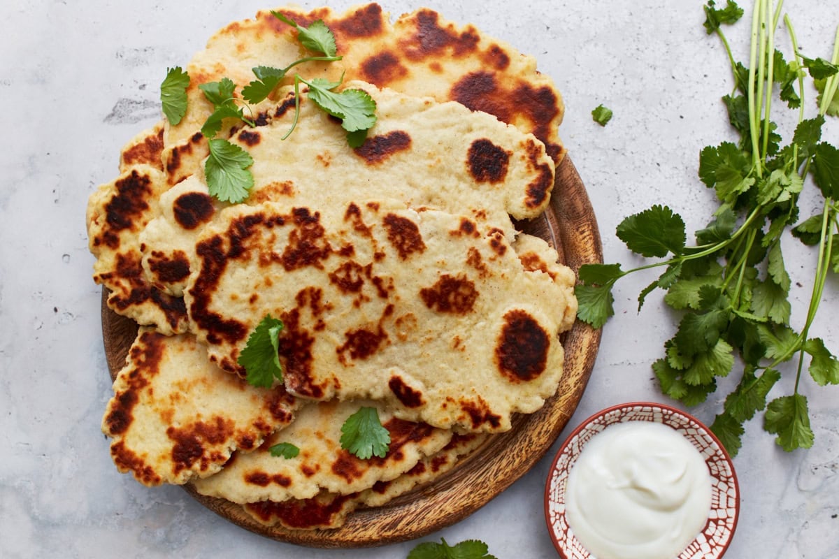 A plate of golden-brown gluten-free naan bread garnished with cilantro, served with a side of creamy yogurt sauce.