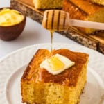 A slice of golden gluten-free sweet cornbread on a white plate, topped with a pat of butter and a drizzle of honey flowing from a honey dipper, with more cornbread visible in the background.