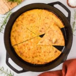 A golden-brown socca flatbread baked in a cast iron skillet, cut into triangular slices, garnished with fresh rosemary and grated cheese.