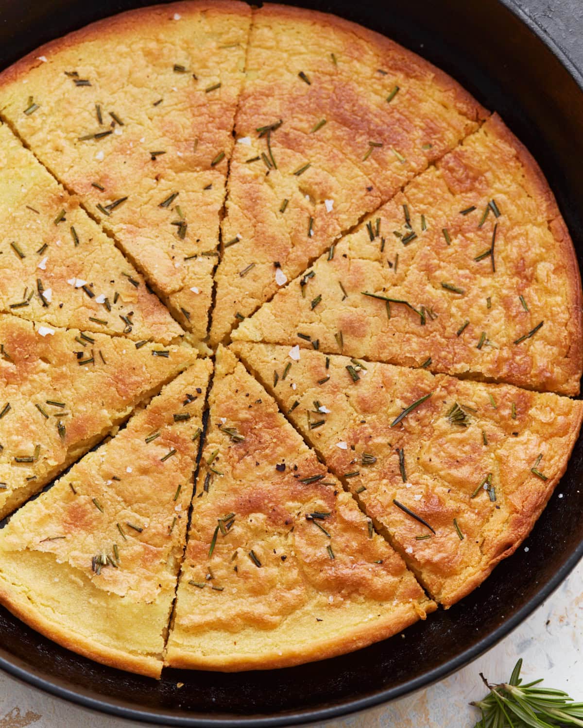 A top view of a whole farinata in a cast iron skillet, cut into wedges and garnished with rosemary and flaky salt.