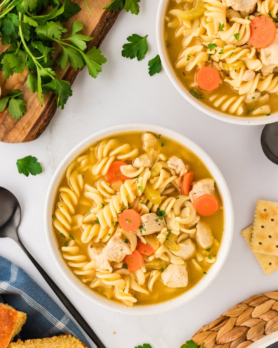 Two bowls of gluten-free chicken noodle soup with spiral noodles, chicken, and vegetables, surrounded by crackers, parsley, and a blue kitchen towel.