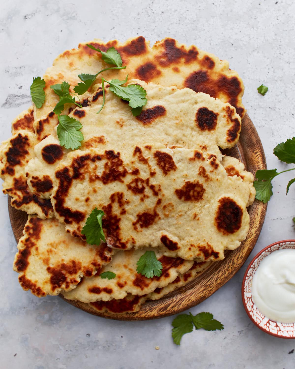 Overhead view of multiple pieces of gluten-free naan bread stacked on a wooden plate, surrounded by cilantro leaves and a bowl of yogurt sauce.
