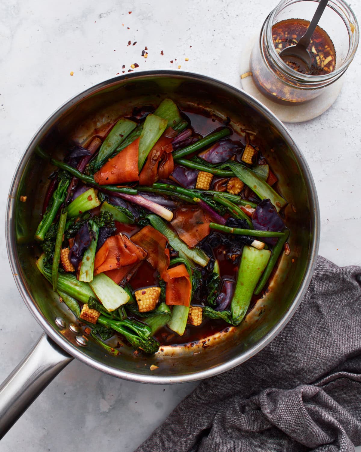 A sauté pan filled with colorful stir-fried vegetables, including bok choy, baby corn, carrots, and purple cabbage, tossed in a glossy gluten-free stir-fry sauce. A jar of the sauce sits nearby.