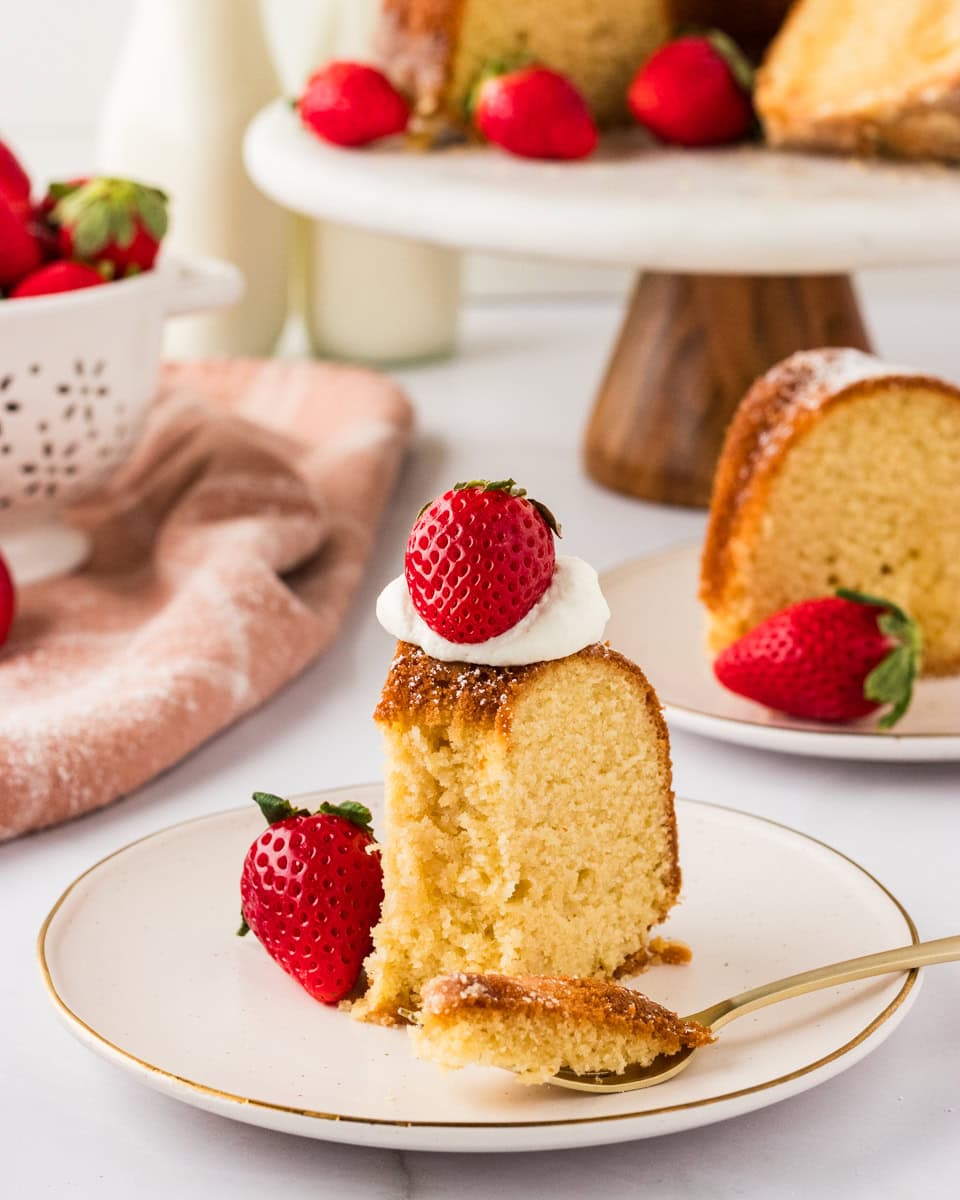 A close-up of a slice of Gluten-Free Kentucky Butter Pound Cake on a plate with a forkful of cake next to it. Fresh strawberries and the whole cake are visible in the background.
