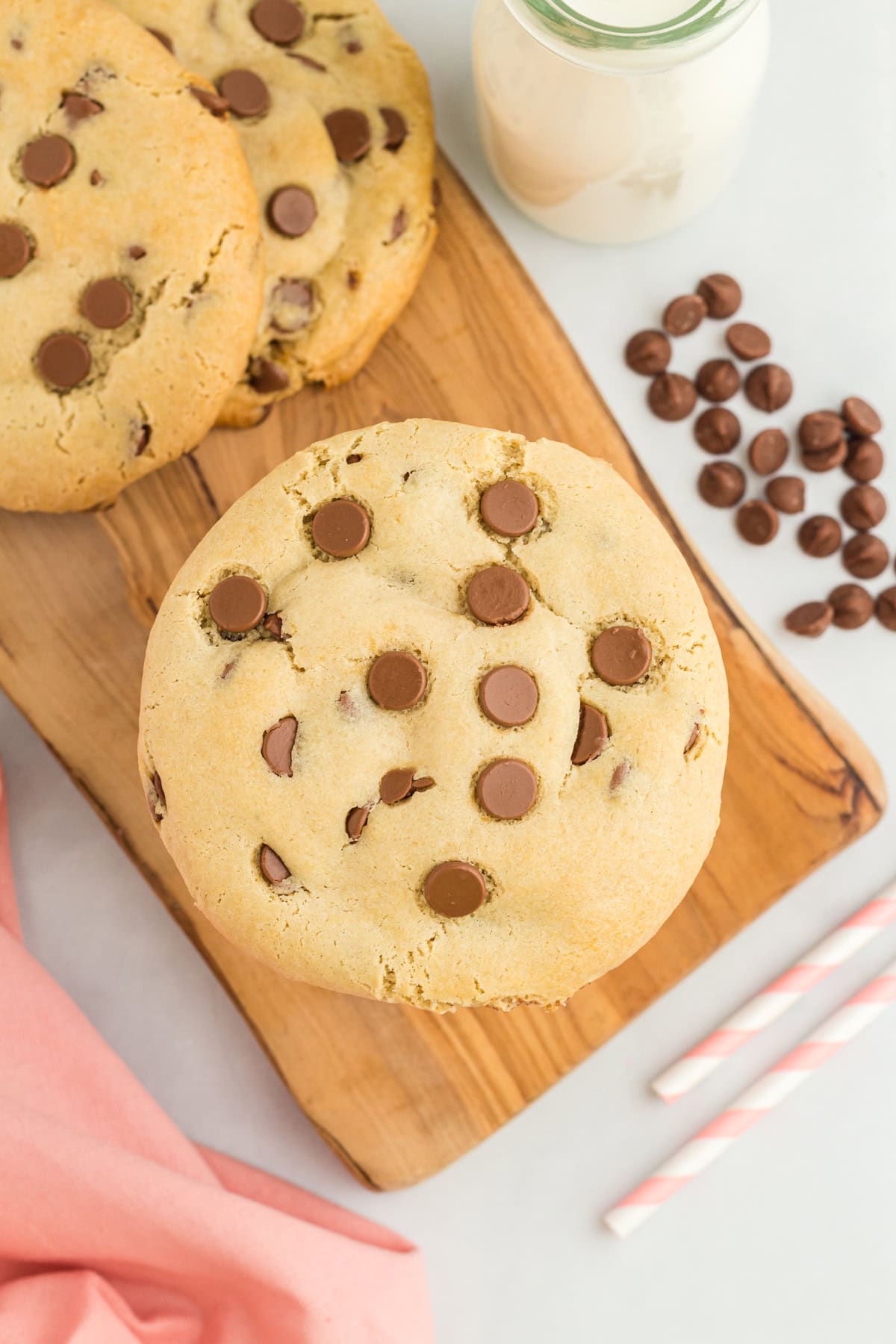 Gluten-free Crumbl chocolate chip cookies stacked on a wooden board, accompanied by scattered chocolate chips, a pink napkin, striped straws, and a glass bottle of milk in the background.