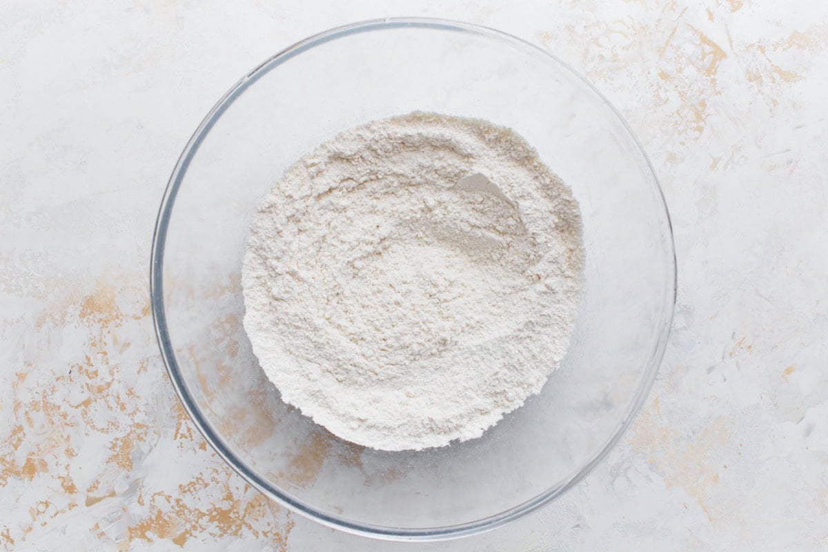 A glass mixing bowl filled with sifted gluten-free flour mixture for the shortbread base, set on a lightly textured surface.
