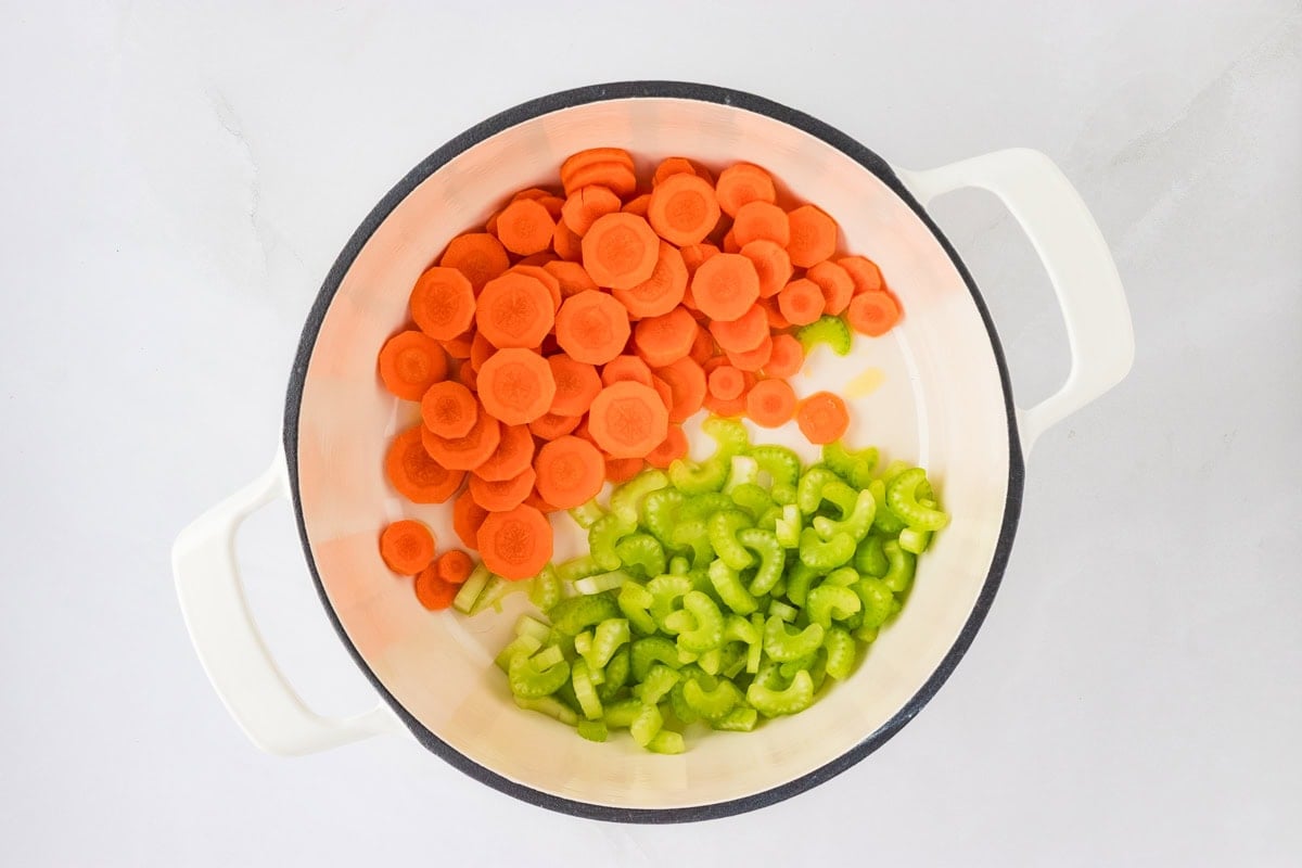 A white pot filled with sliced raw carrots and celery, ready for cooking, against a light background.