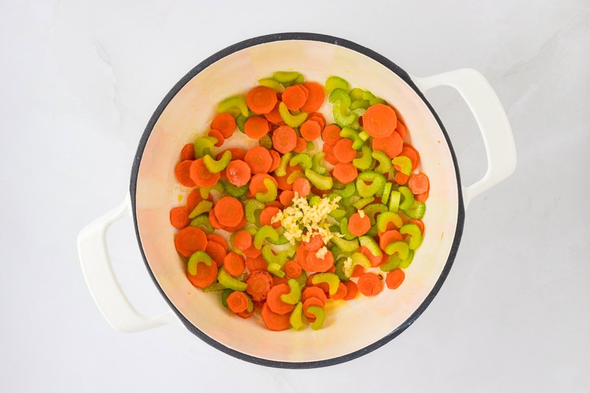 A white pot with sautéed carrots, celery, and minced garlic, showcasing the preparation steps for the soup.