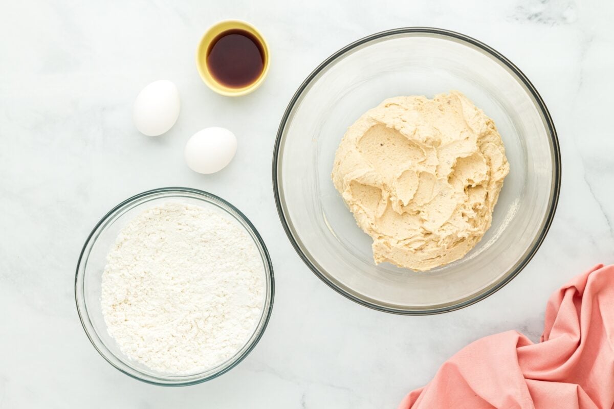 Ingredients laid out for the recipe, including two eggs, a bowl of vanilla extract, a bowl of flour, and a bowl of creamed butter and sugar mixture.