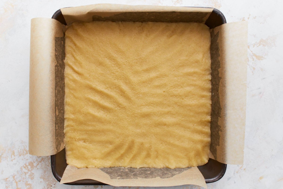 A baking pan lined with parchment paper, filled with evenly spread gluten-free shortbread dough before baking.