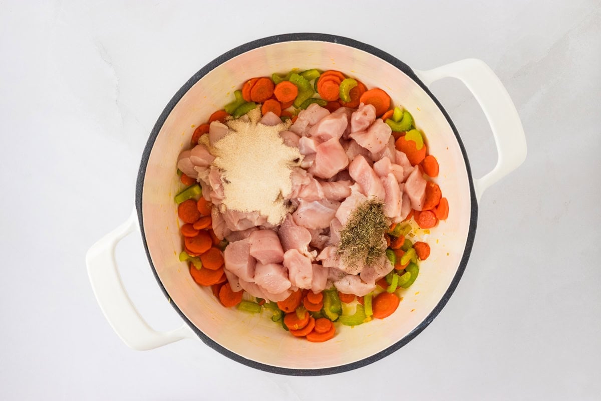 A white pot with raw chicken chunks, sautéed vegetables, and seasonings like dried herbs, showcasing the ingredients.