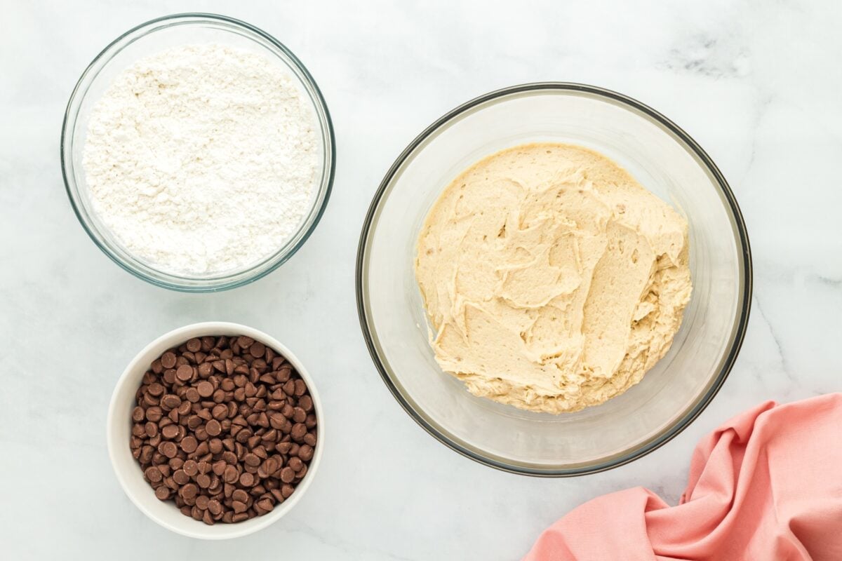 A mixing setup showing three bowls: one with gluten-free flour, one with creamed butter and sugar, and another with chocolate chips, ready to be combined.