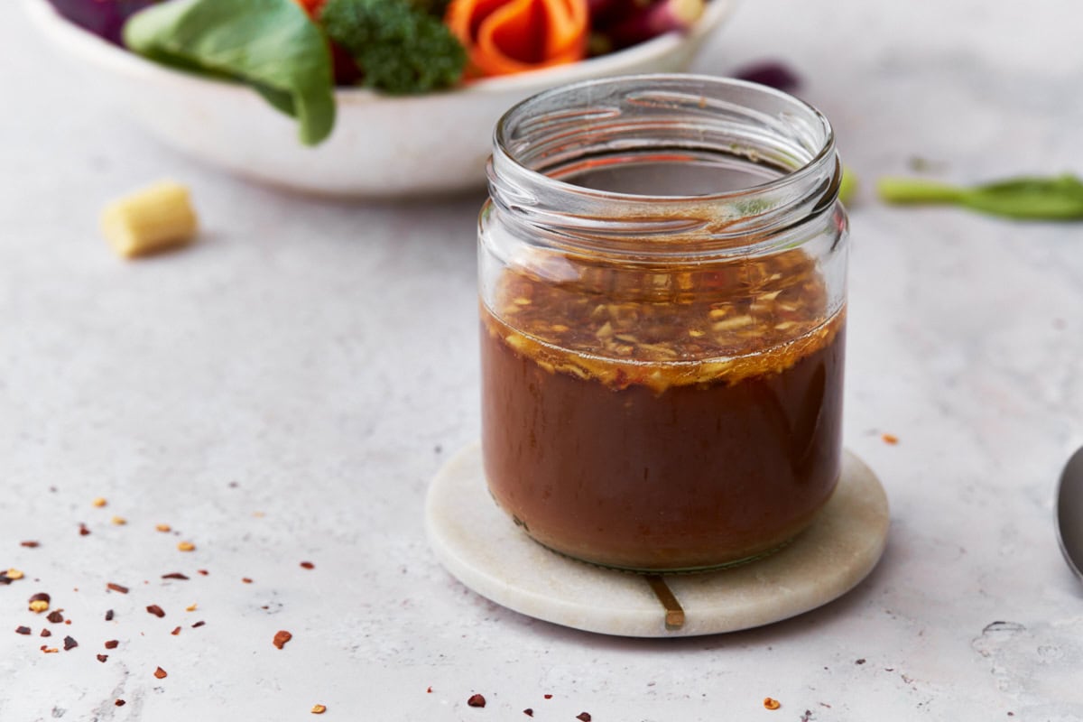 A jar of gluten-free stir-fry sauce placed on a white coaster, with a blurred bowl of fresh vegetables, including broccoli and carrots, in the background.