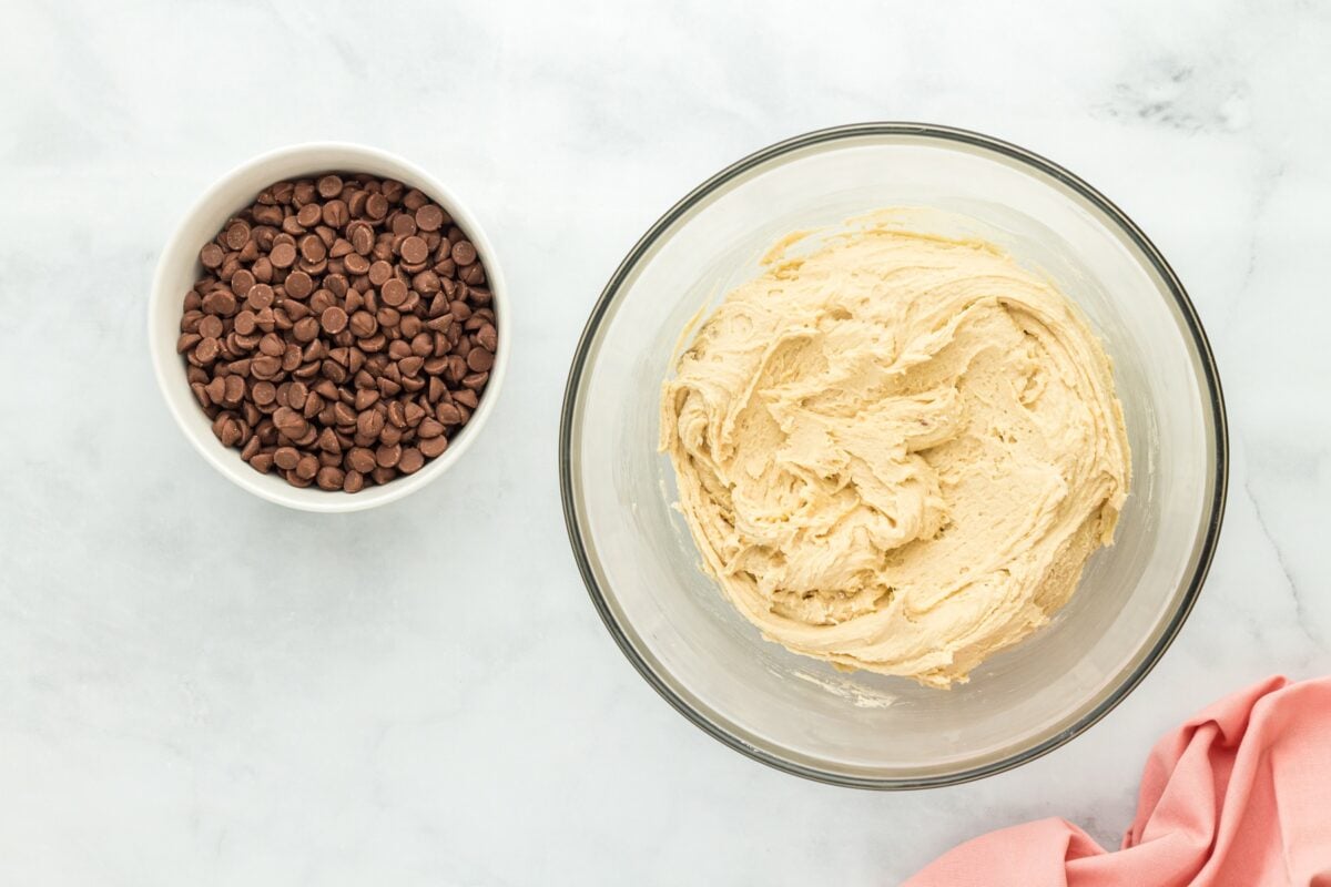 A mixing bowl filled with smooth creamed butter and sugar mixture, alongside a separate bowl of chocolate chips.