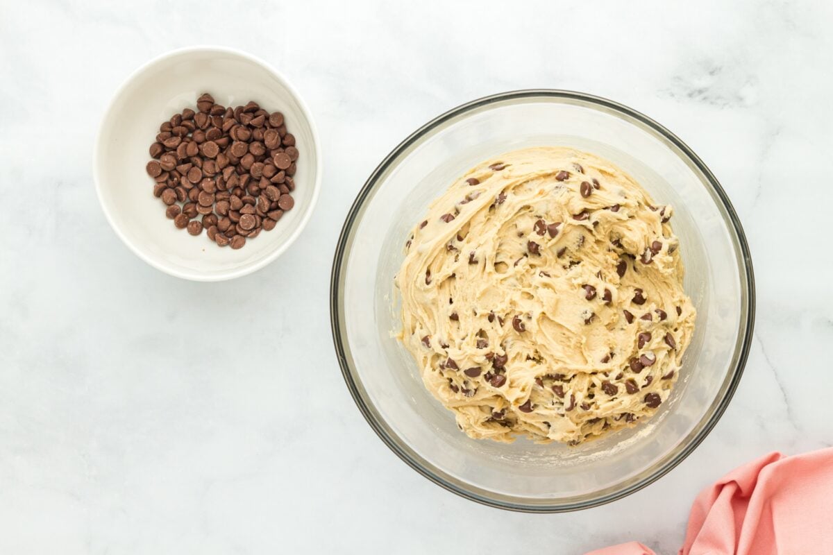 A large mixing bowl containing cookie dough mixed with chocolate chips, next to a bowl of extra chocolate chips for topping.