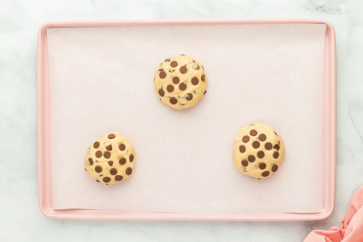 Three unbaked gluten-free cookie dough balls, topped with chocolate chips, spaced out on a parchment-lined pink baking sheet.