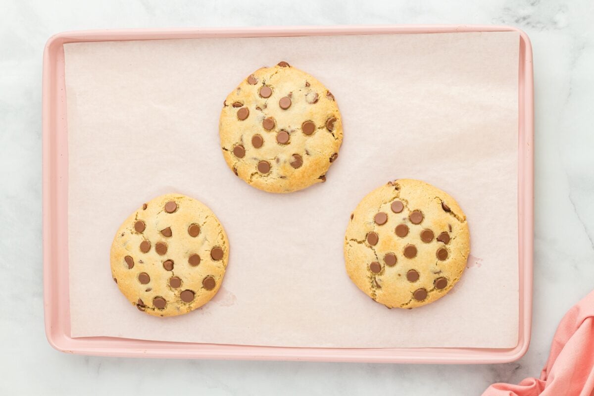 Three freshly baked gluten-free Crumbl chocolate chip cookies on a parchment-lined pink baking sheet, golden brown and topped with melted chocolate chips.