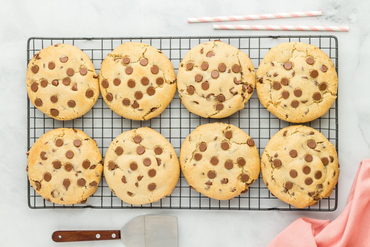 A cooling rack holding eight large gluten-free Crumbl chocolate chip cookies, golden brown with chocolate chips evenly distributed, with pink-striped straws in the background.