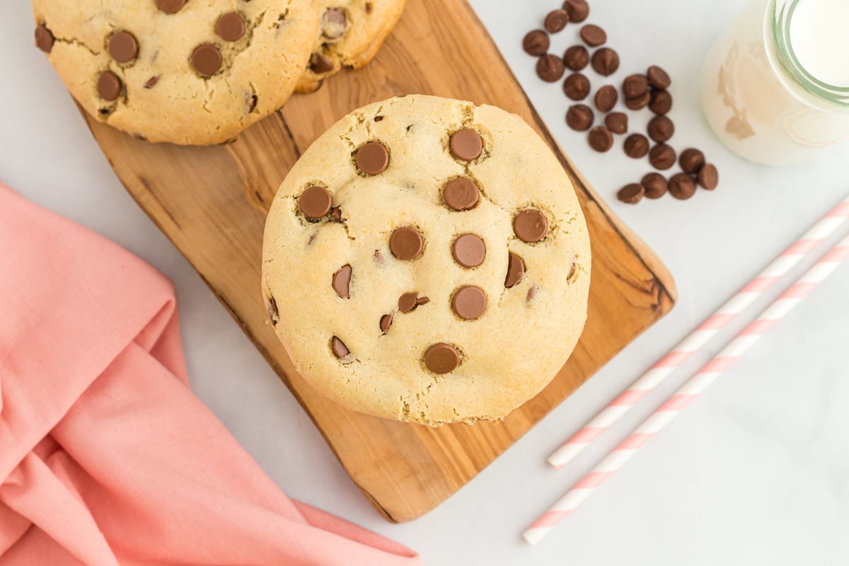 A close-up view of a gluten-free Crumbl chocolate chip cookie placed on a wooden board, surrounded by chocolate chips, a glass of milk, and pink-striped straws.

