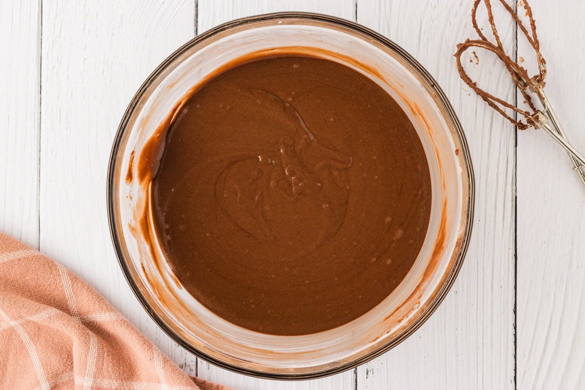 The fully mixed, smooth, and glossy chocolate cake batter in a glass mixing bowl, with a hand mixer coated in batter resting beside it.