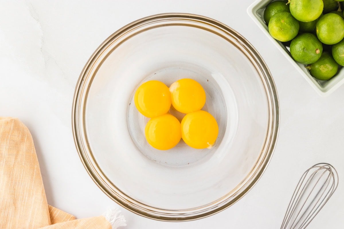 xA glass mixing bowl with four separated egg yolks, ready to be whisked for the key lime pie filling, placed next to fresh limes and a wire whisk.