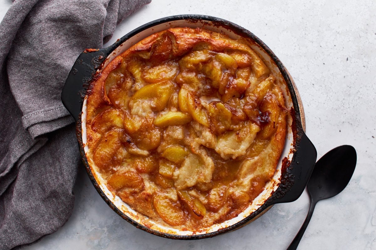 A baked peach cobbler in a black skillet placed on a surface next to a gray napkin and a black serving spoon.