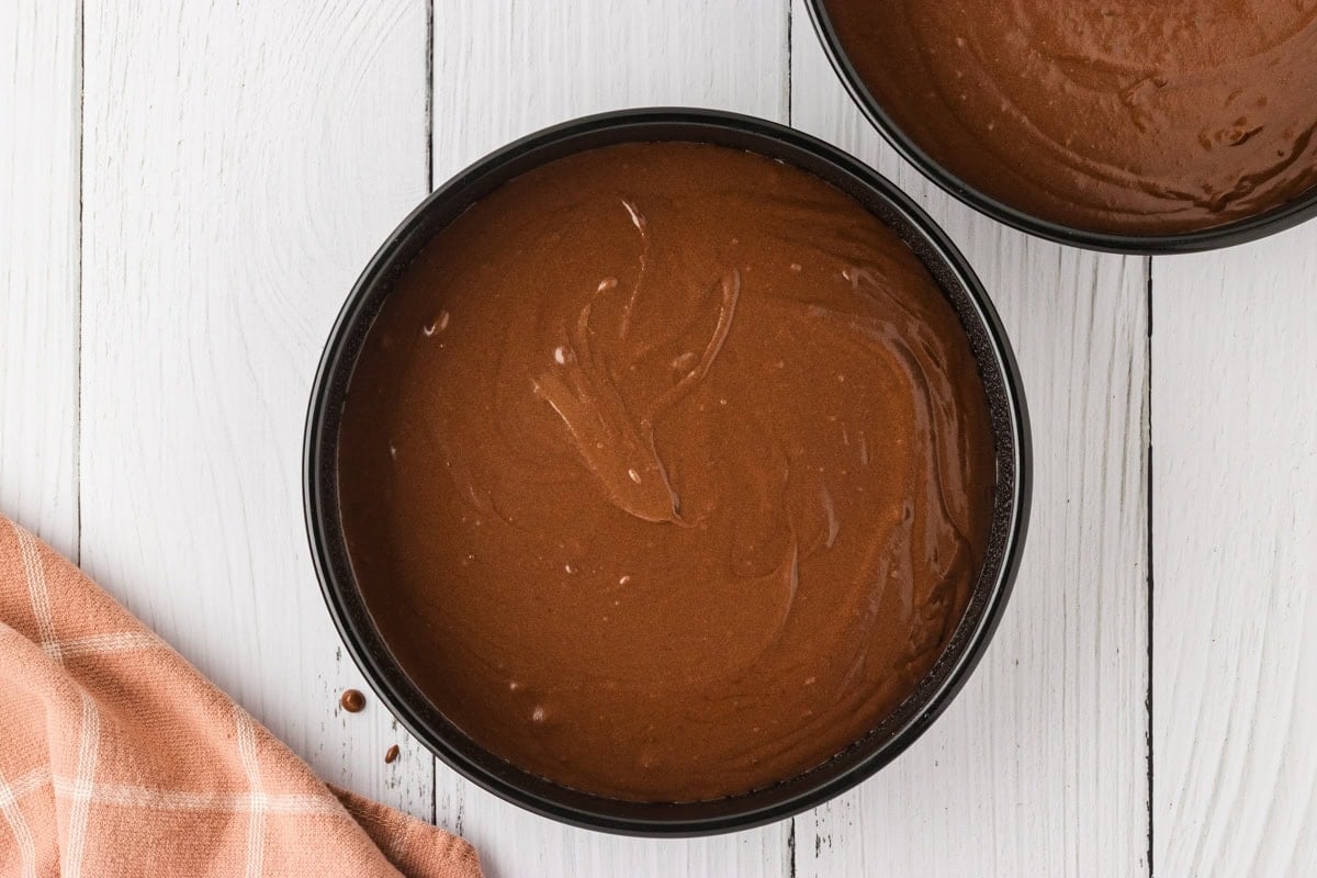 Two round cake pans filled with smooth gluten-free chocolate cake batter, ready for baking, placed on a white wooden surface.