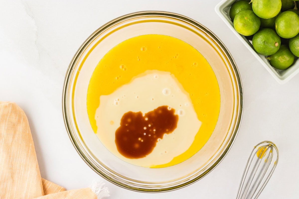 A glass bowl with whisked egg yolks, sweetened condensed milk, and vanilla extract, ready to be mixed for the key lime pie filling. Fresh limes and a whisk sit nearby.