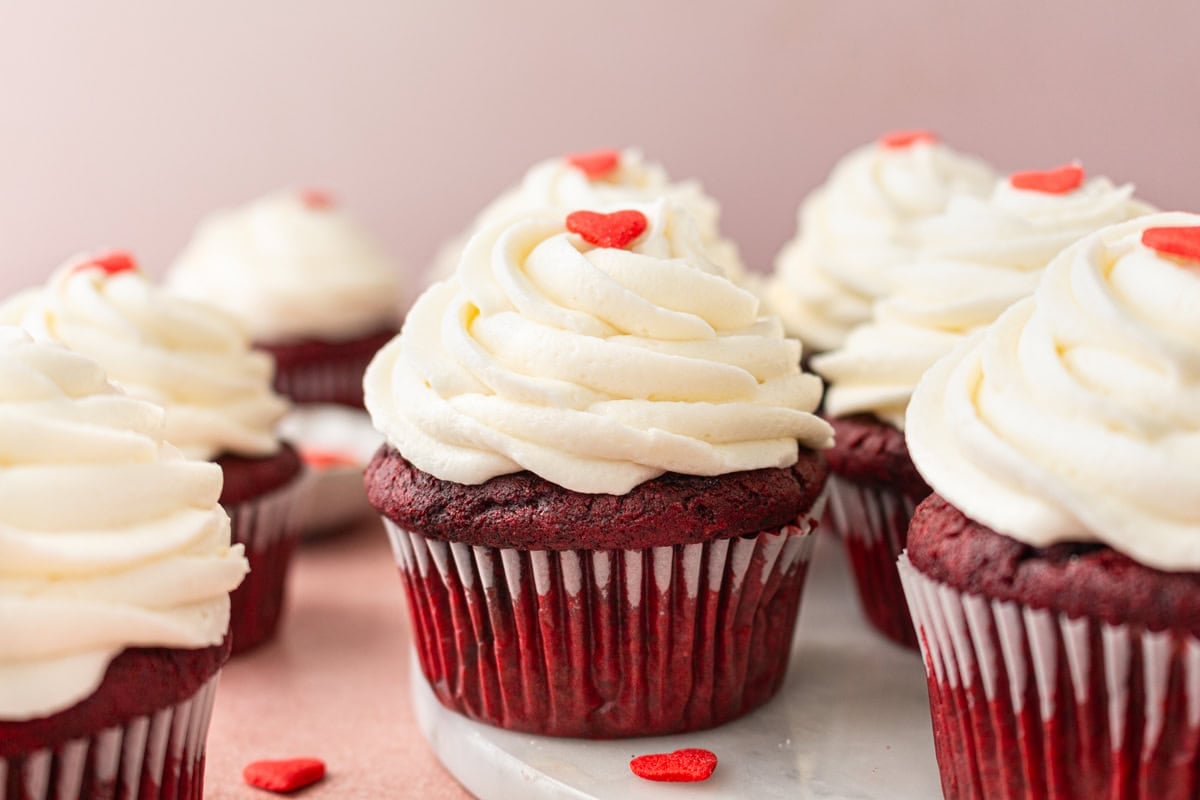Close-up of gluten-free red velvet cupcakes on a marble serving board, with fluffy cream cheese frosting piped in elegant swirls and red heart sprinkles for decoration.