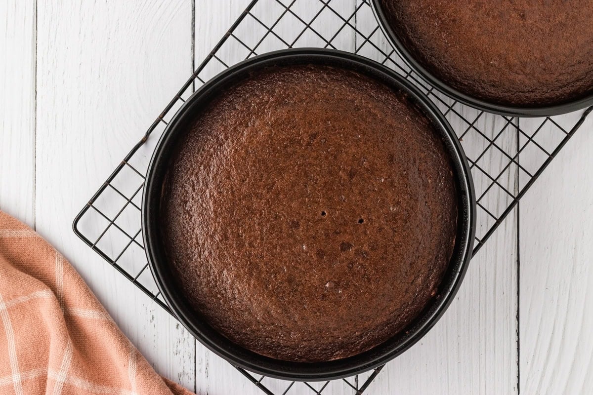 Two freshly baked gluten-free chocolate cake layers in round cake pans, cooling on a black wire rack.