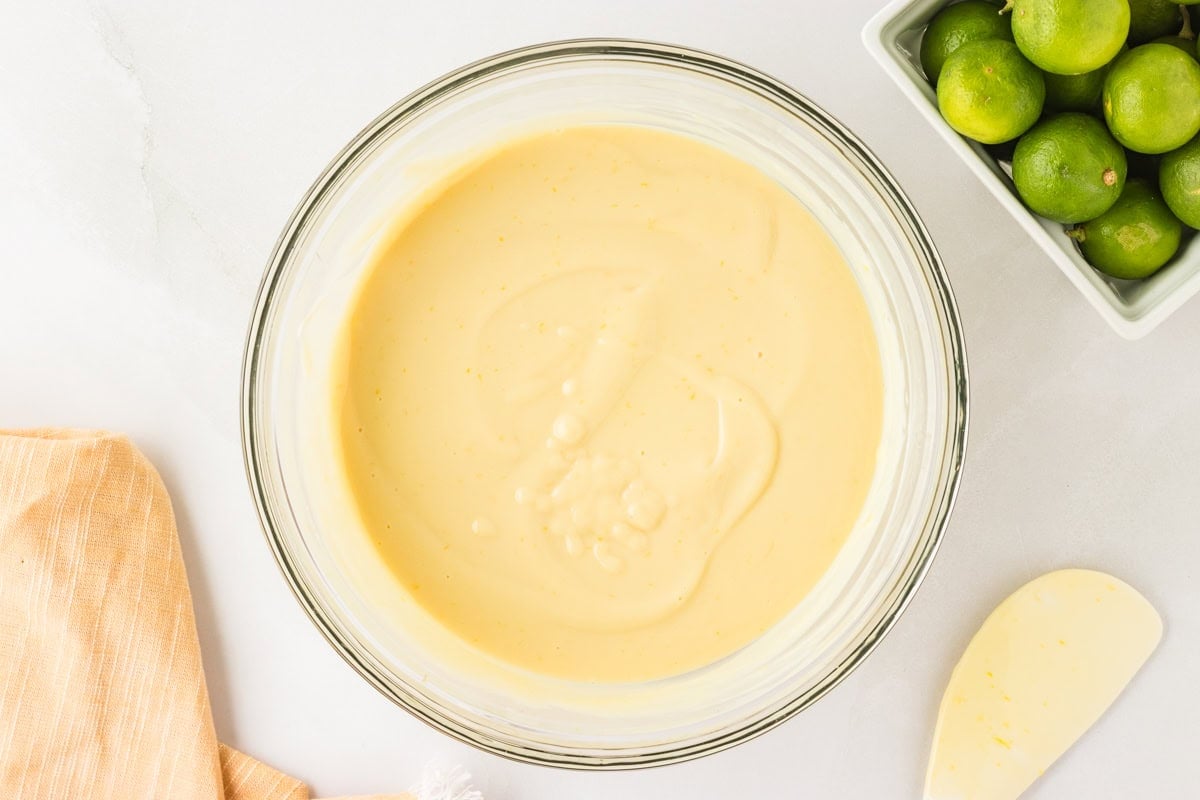 A bowl of fully mixed, creamy key lime filling with a pale yellow color, placed on a white countertop next to a spatula and fresh limes.