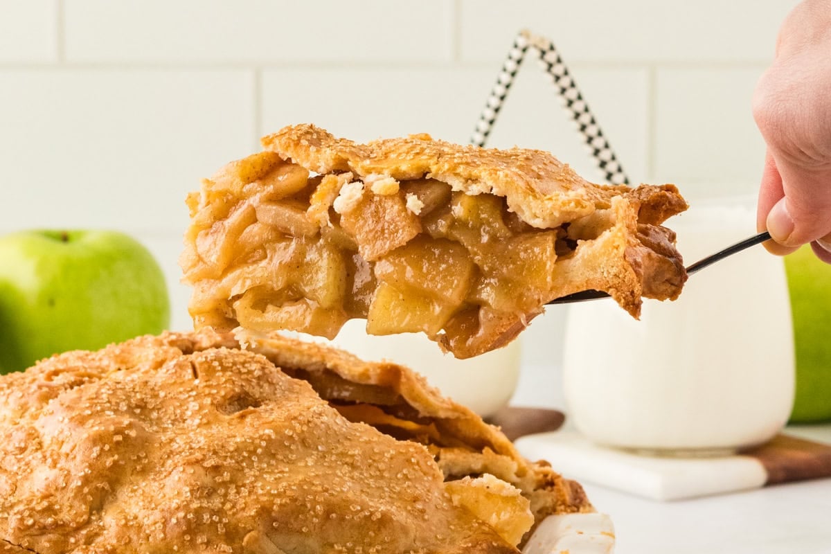 A freshly baked gluten-free apple pie with a crisp, golden crust is being sliced, revealing a thick apple filling. A hand holds a server lifting a piece, while whole green apples and glasses of milk sit in the background.