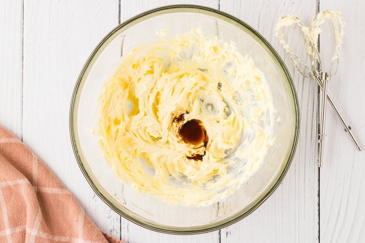 A glass bowl with whipped butter and vanilla extract, ready to be mixed for frosting, with a hand mixer beside it.