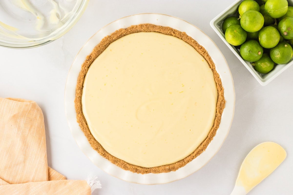 A white pie dish filled with smooth gluten-free key lime pie filling inside a gluten-free Chex crust, ready for baking. A spatula with excess filling is placed nearby.