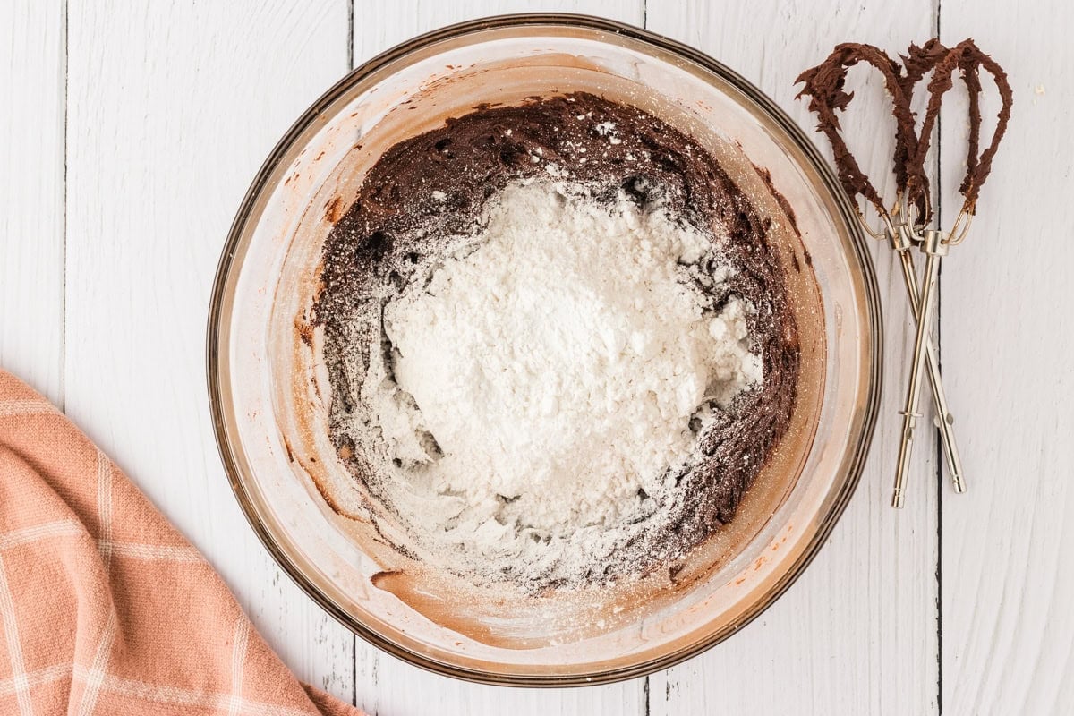 A glass bowl with chocolate frosting, with a mound of powdered sugar added on top, ready to be mixed.
