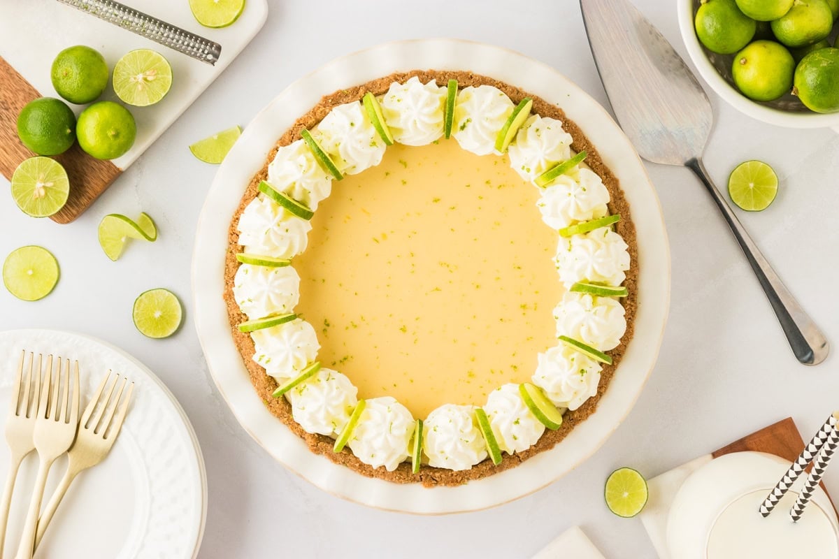A fully decorated gluten-free key lime pie with whipped cream rosettes and thin lime slices arranged around the edges, dusted with lime zest, ready to be served.